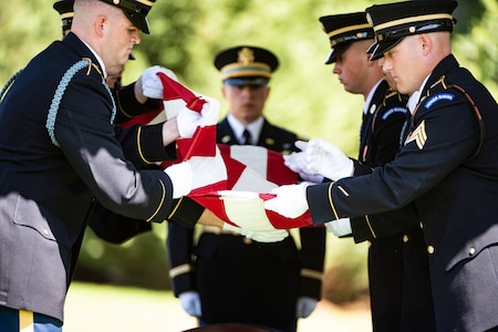 Several Army soldiers in dark ceremonial uniforms and white gloves are leaning towards each other as they fold a US flag over a brown casket.