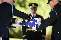 Army soldiers wearing dark ceremonial uniforms and white gloves are folding a US flag while another looks on.