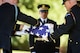 Army soldiers wearing dark ceremonial uniforms and white gloves are folding a US flag while another looks on.