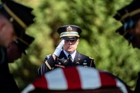 A military member dressed in dark ceremonial uniform with white gloves is saluting towards a US Flag-draped casket as other members make adjustments to the flag.