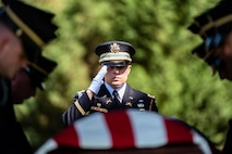 A military member dressed in dark ceremonial uniform with white gloves is saluting towards a US Flag-draped casket as other members make adjustments to the flag.