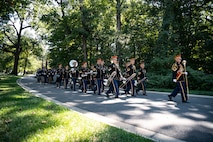 Military members in dark ceremonial uniforms with red round hats are marching down a winding street with lots of trees in the background. Most are carrying musical instruments