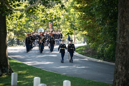 Military members dressed in dark ceremonial uniforms are marching down a winding road. There are two in the front leading a larger group who are marching in three columns, carrying musical instruments. There are a few white tombstones in the foreground surrounded by green grass.