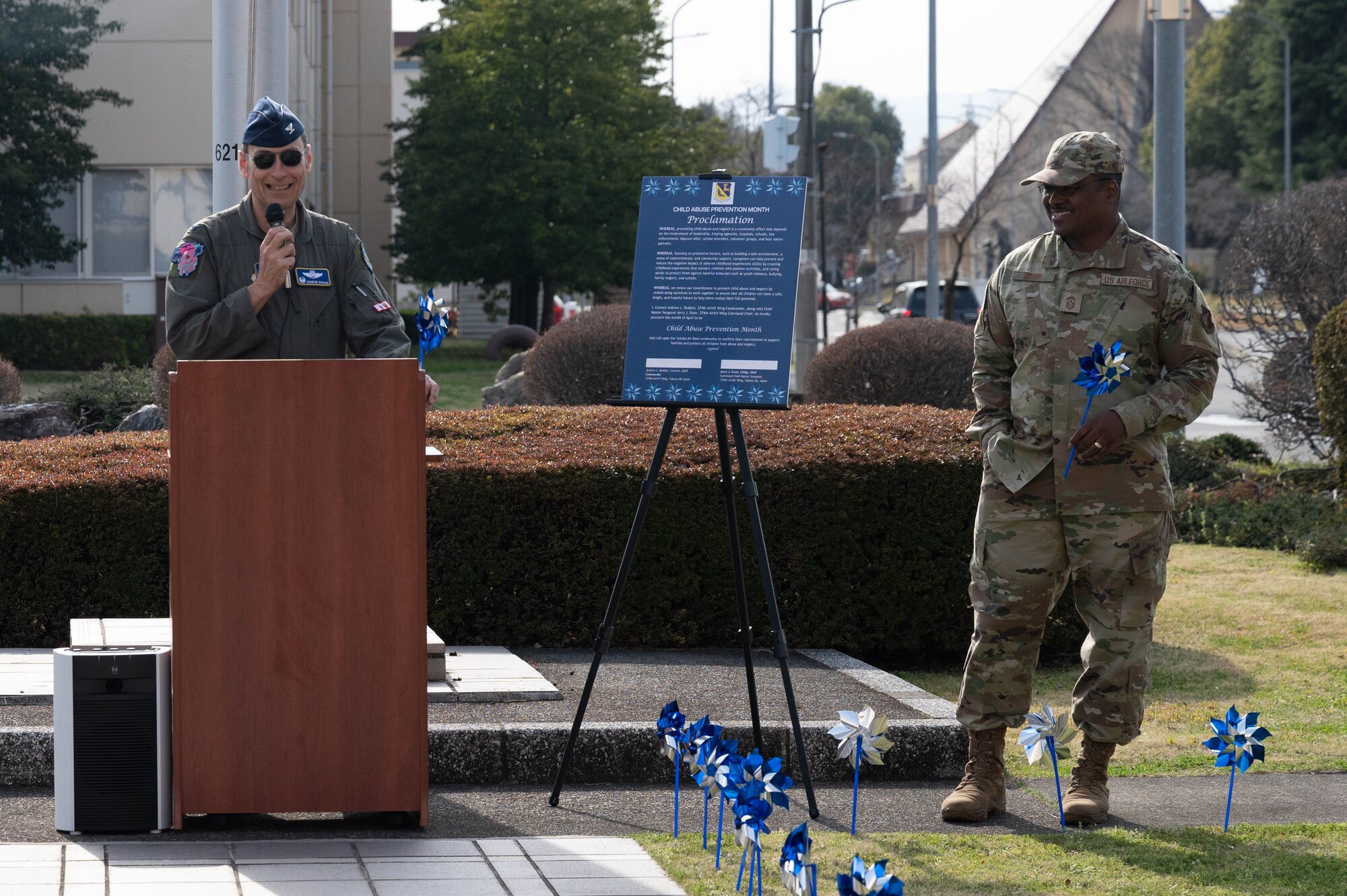 An Airman speaks at a podium and another Airman stands to the side while holding a pinwheel.