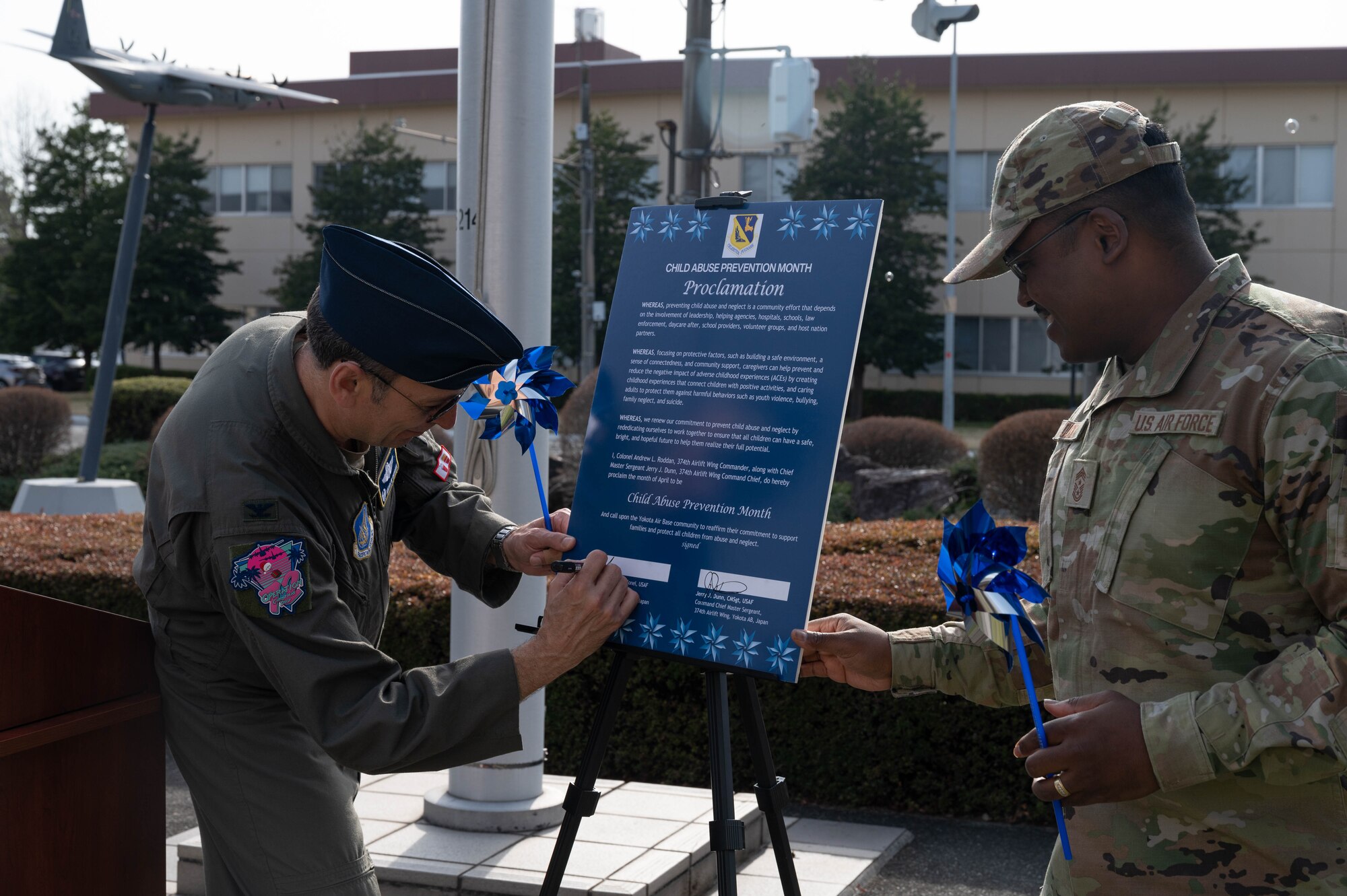 An Airman signs a proclamation and another Airman stands while holding a pinwheel.
