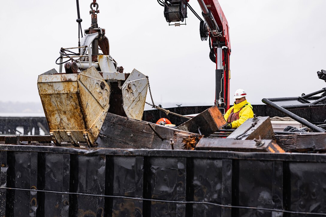A person wearing a yellow jacket and white hard hat stands by an excavator amid metal debris.