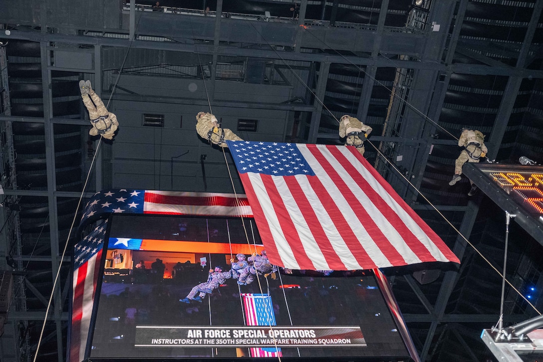 Four airmen use cables to descend from the top of an arena with a large American flag in the middle of them and a large video screen behind them.