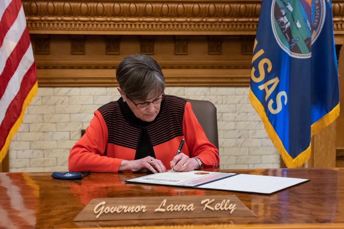 Governor Kelly sits at her desk and signs a bill.
