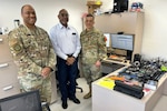 Two Soldiers in Uniform and a University Professor pose for a group shot behind a desk, with computer parts and cables