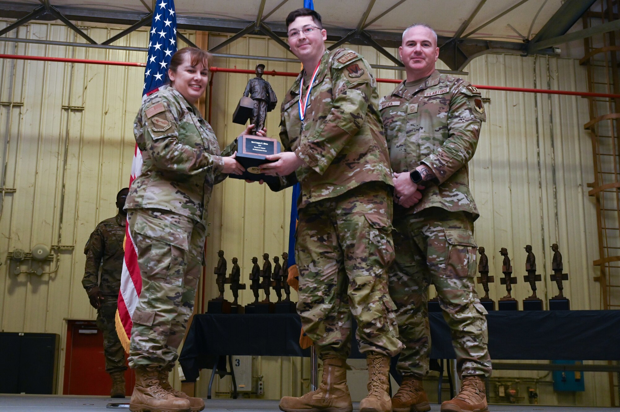 Senior Airman Logan Brice, center, assigned to the 333th Fighter Generation Squadron, receives the Chief Master Sgt. Thomas N. Barnes award from Col. Kathryn Roman, 4th Maintenance Group commander and Chief Master Sgt. Michael Bylica, 4 MXG senior enlisted leader, during the 4 MXG Maintenance Person of the Year ceremony at Seymour Johnson Air Force Base, North Carolina, Mar. 08, 2024. The 4th Maintenance Group hosts the event annually for MXG Airmen to celebrate their achievements and hard work. (U.S. Air Force photo by Airman Rebecca Tierney)