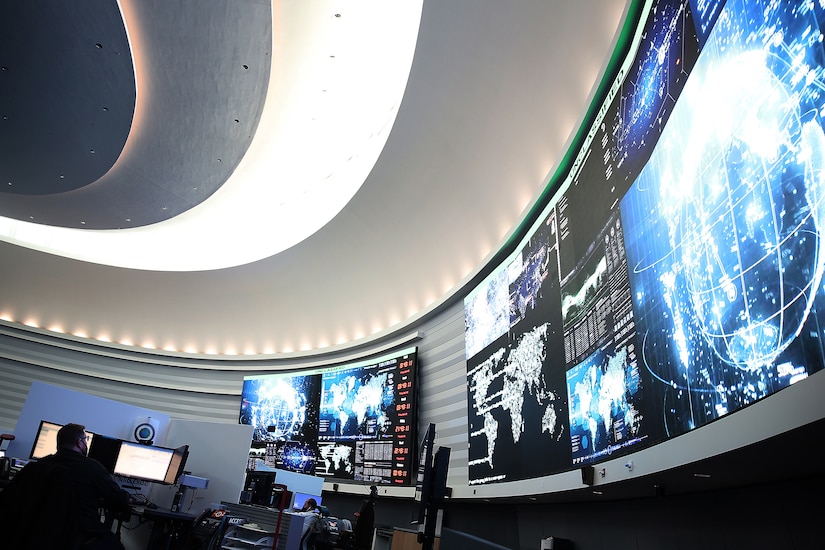 A man in civilian attire sits at a computer terminal on a watch floor.