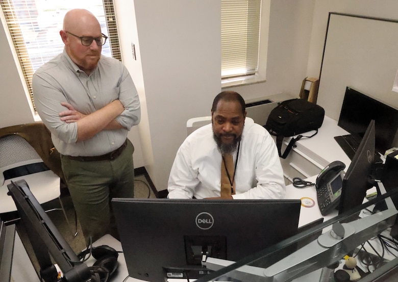 Man standing at a desk overlooking another man sitting down looking at computer.