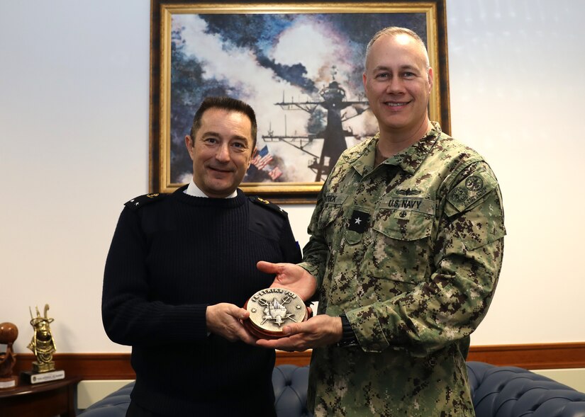 A French sailor and an American sailor hold a bronze token with a painting of a ship in the backdrop.