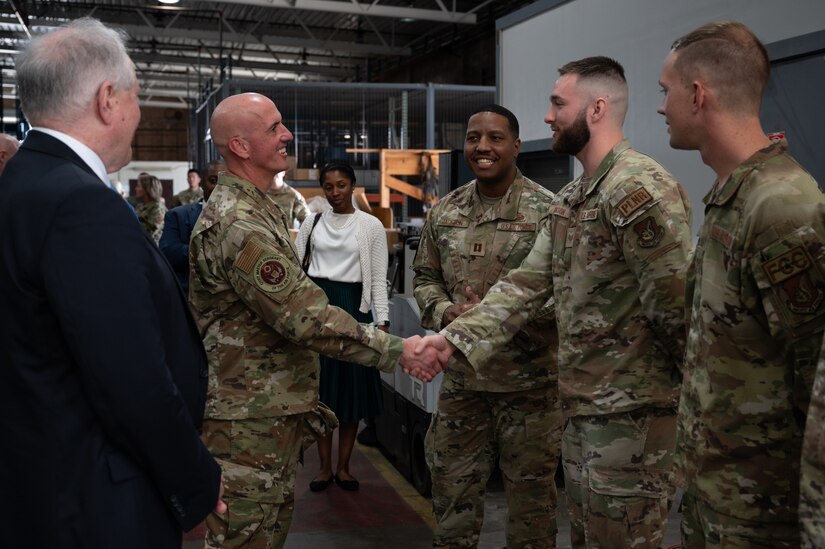Two airmen shake hands while others stand around in a facility with exposed beams