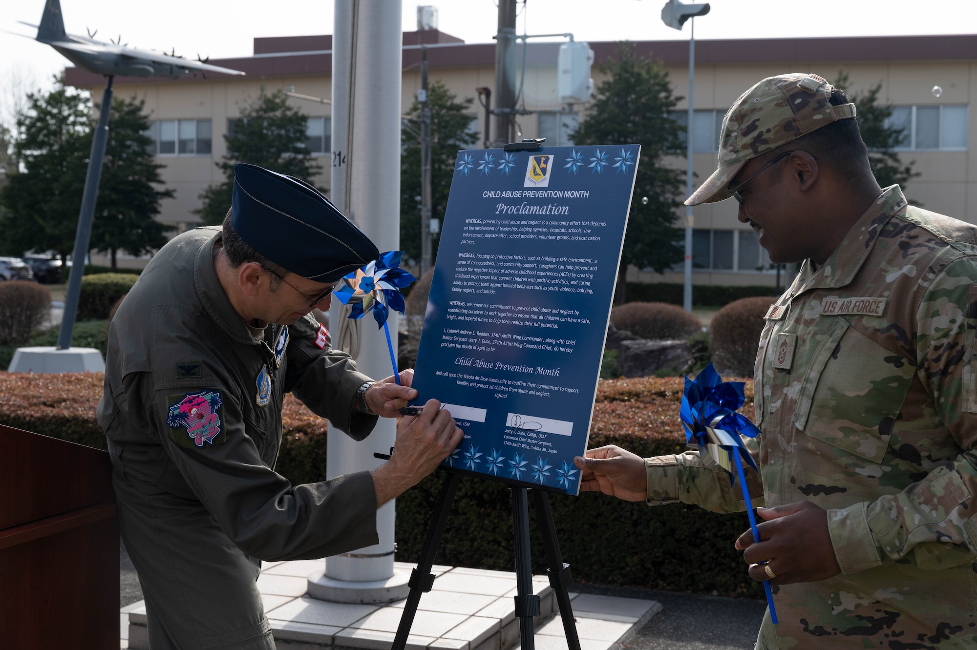 An Airman signs a proclamation and another Airman stands while holding a pinwheel.