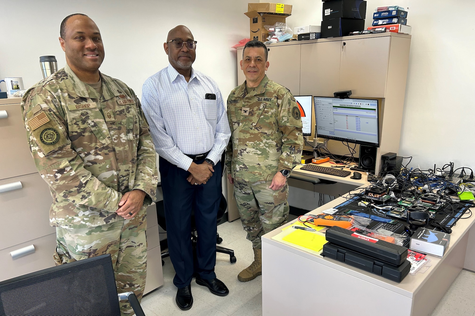 Two Soldiers in Uniform and a University Professor pose for a group shot behind a desk, with computer parts and cables