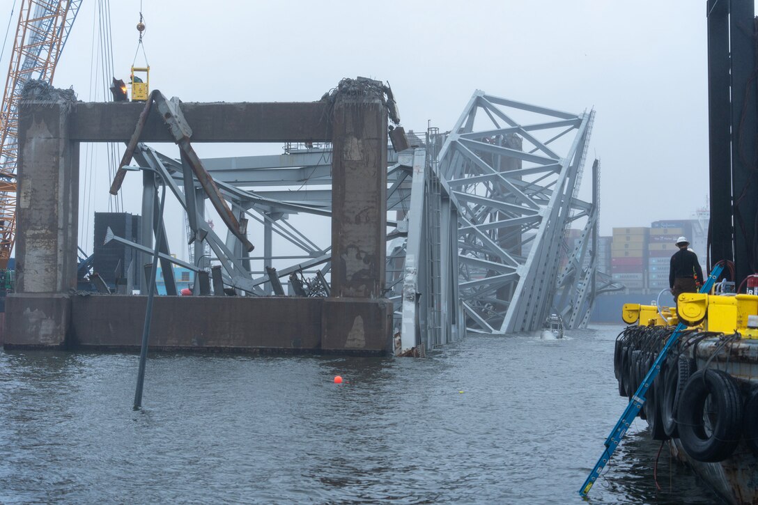A worker in a bucket raised by construction equipment torches an area on a collapsed metal bridge that is partly submerged in water.