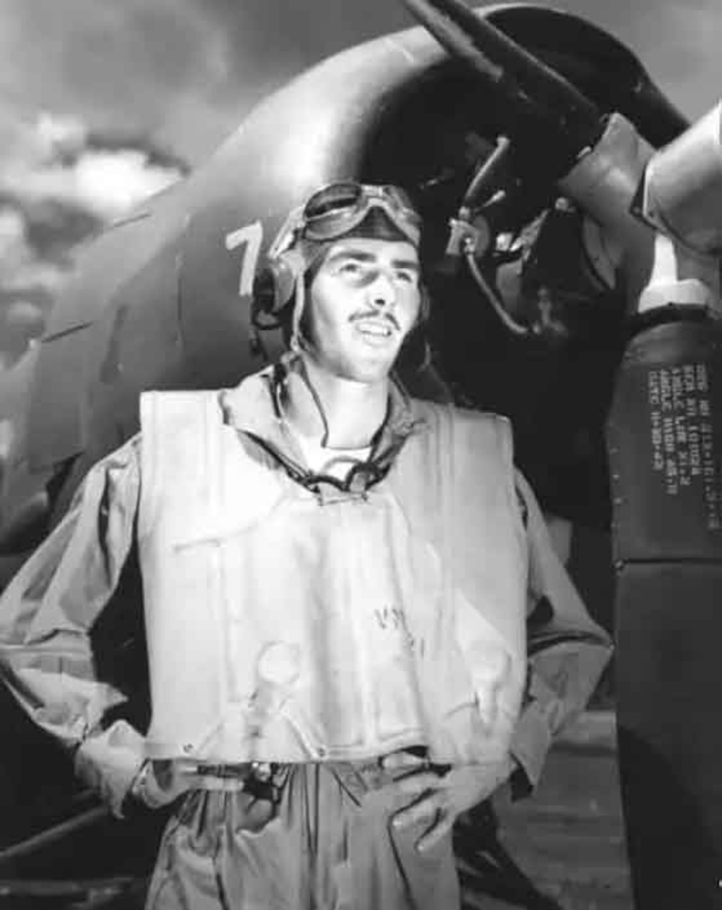 A man in flight suit stands with his hands on his hips in front of an airplane.
