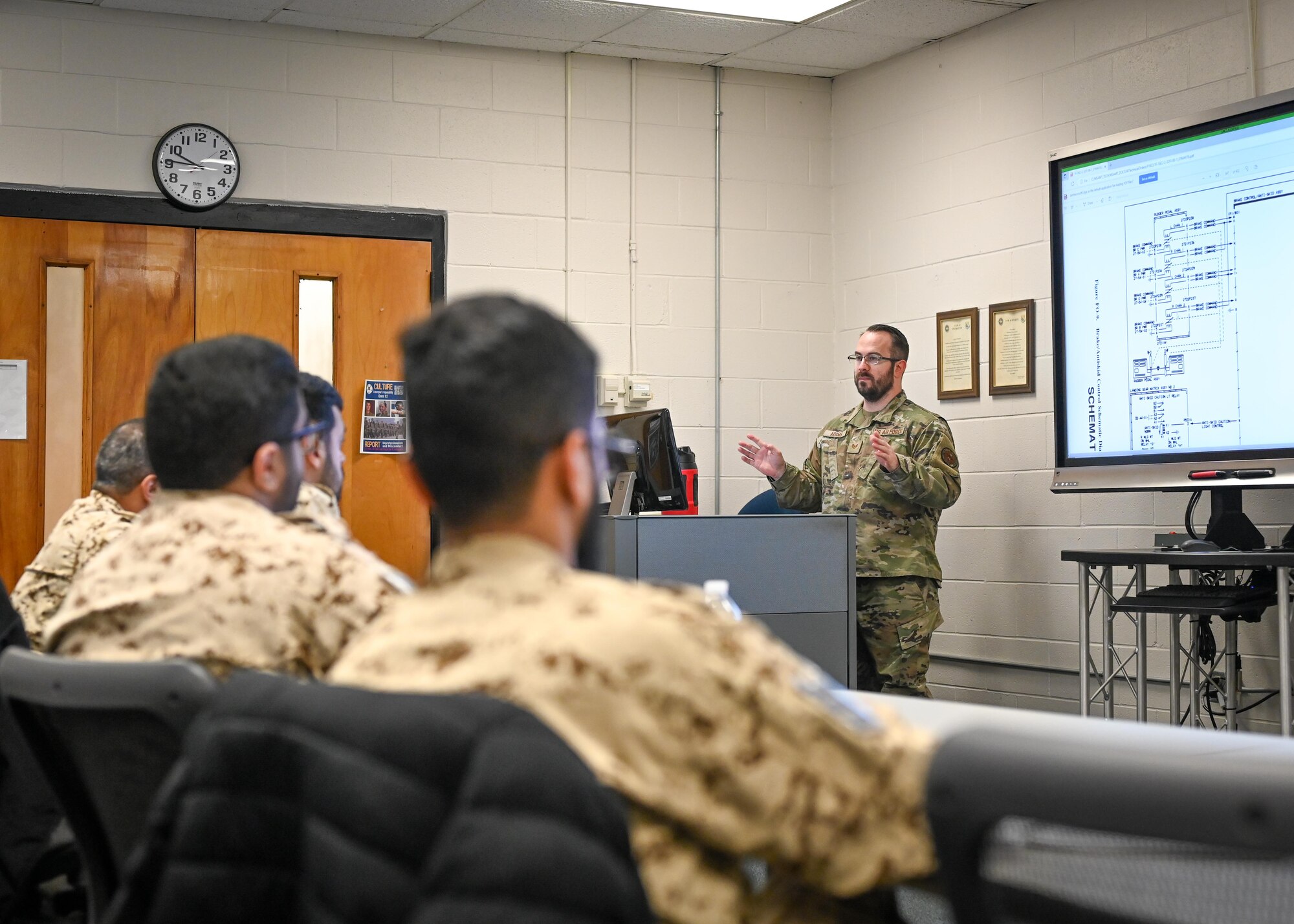 A man presents in front of a screen to a group of men sitting.