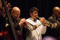 A young boy with dark hair is wearing a white suit with a black bowtie, and his is playing on a brass trumpet next to men in dark Army ceremonial uniforms who are also playing instruments.