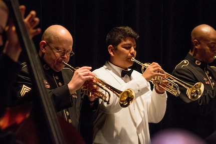 A young boy with dark hair is wearing a white suit with a black bowtie, and his is playing on a brass trumpet next to men in dark Army ceremonial uniforms who are also playing instruments.
