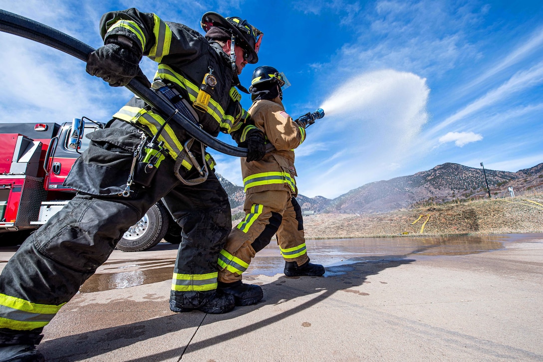 Two people in firefighting gear and a fire truck in the background aim a hose spraying water toward mountains in the distance.