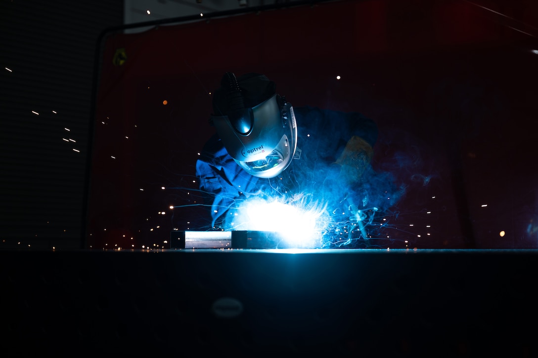 An airman wearing a helmet and visor in a blue light creates bright sparks while welding metal in a dark room.