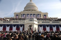 Crowds of people in winter weather clothing are gathered in front of the U.S. Capitol building. There are red, white and blue flags and banners draped on many parts of the building, and members of the military dressed in various uniforms are facing the crowd. One military member is conducting a band.