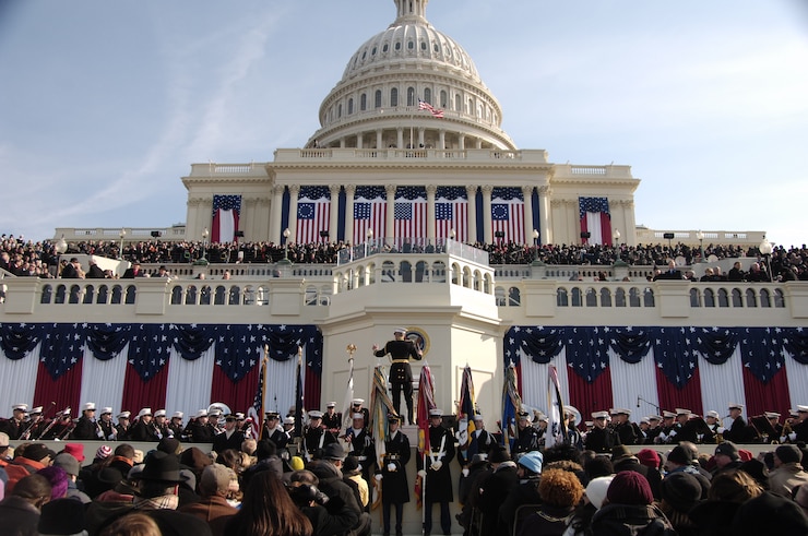 Crowds of people in winter weather clothing are gathered in front of the U.S. Capitol building. There are red, white and blue flags and banners draped on many parts of the building, and members of the military dressed in various uniforms are facing the crowd. One military member is conducting a band.