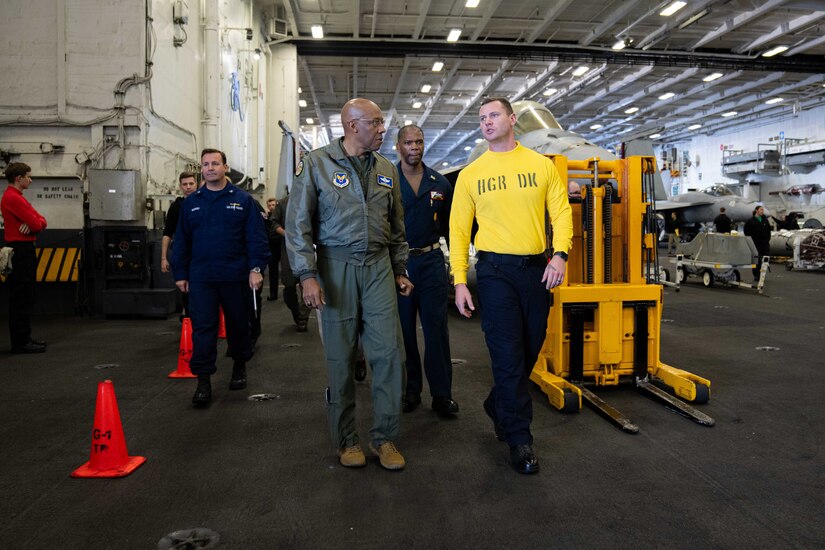 A man in a flight suit walks next to another man inside the hangar bay of an aircraft carrier. In the background are three men in coveralls and a forklift.