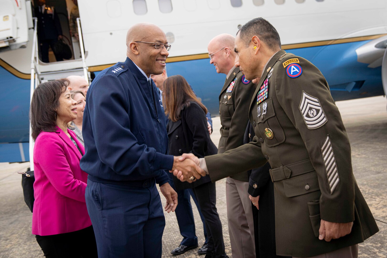 A man in uniform shakes hands with a soldier. A jet airplane with the stairway down is behind them.