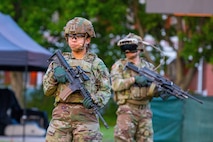 Two soldiers wearing green Army camouflage combat uniforms with helmets, and one with black goggles on, are looking around while holding black rifles in front of them. They are on a green lawn.