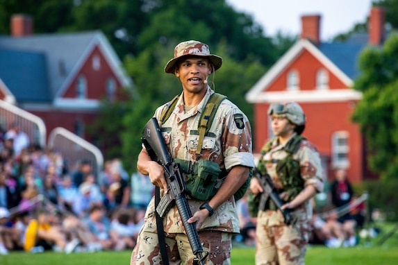 A soldier dressed in Desert Storm/Desert Shield-era Army green uniform with cap with full brim while carrying a rifle and speaking to a crowd who is seen in the background seated. He's on a green lawn.