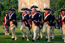 Soldiers dressed in blue with red trim and white pants Revolutionary War-ear uniforms are charging towards the camera at an angle with rifles and bayonets affixed in front of them. They are on a green lawn