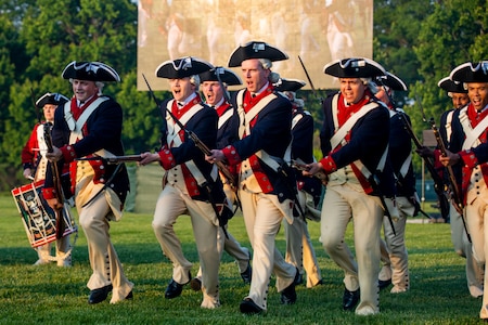 Soldiers dressed in blue with red trim and white pants Revolutionary War-ear uniforms are charging towards the camera at an angle with rifles and bayonets affixed in front of them. They are on a green lawn