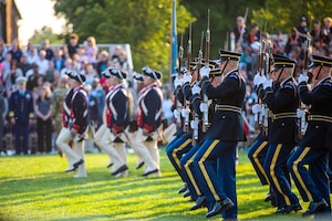 Soldiers dressed in dark ceremonial uniforms holding brown rifles in front of them are marching forward (to the left in the picture) in the foreground. There are soldiers dressed in dark blue Revolutionary War-era uniforms marching forward in the same direction in the background. They are on a green lawn.