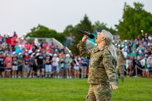 A woman with long blond air tied back into a ponytail is wearing a green Army camouflage uniform while holding a microphone and singing upwardly. She's on a green lawn with a large crowd in the background on bleachers.