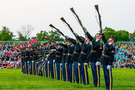 Army soldiers dressed in dark ceremonial uniforms are standing in a straight row on a green lawn while lifting brown rifles into the air one by one which is forming the look of a wave.