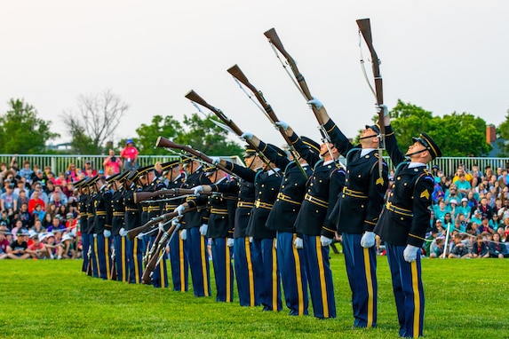 Army soldiers dressed in dark ceremonial uniforms are standing in a straight row on a green lawn while lifting brown rifles into the air one by one which is forming the look of a wave.