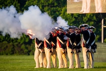 Soldiers dressed in dark blue Revolutionary War-era uniforms are firing long brown rifles, and there is white smoke coming from the guns. They are standing on a green lawn.