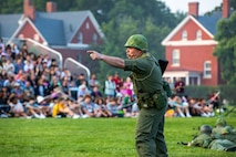A soldier dressed in a Vietnam War-era solid green uniform and helmet is pointing to the left. He is standing on a green lawn in front of a crowd who is seated on bleachers.