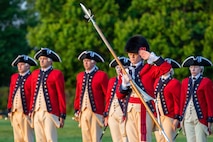 Soldiers dressed in red Revolutionary War-era uniforms are standing rows holding instruments at their sides and the person in front salutes with his palm facing outward while holding a large staff with the a silver spear end.