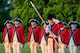 Soldiers dressed in red Revolutionary War-era uniforms are standing rows holding instruments at their sides and the person in front salutes with his palm facing outward while holding a large staff with the a silver spear end.