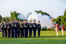 Solders dressed in dark Army ceremonial uniforms are facing towards the right while standing in rows on a green lawn. There is smoke in the background. There are other soldiers dressed in red Revolutionary War-era uniforms on the right side of the picture,