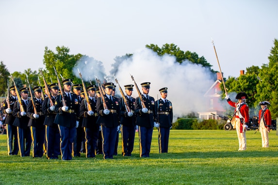 Solders dressed in dark Army ceremonial uniforms are facing towards the right while standing in rows on a green lawn. There is smoke in the background. There are other soldiers dressed in red Revolutionary War-era uniforms on the right side of the picture,