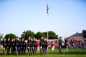 Soldiers in dark ceremonial uniforms are facing to the right while standing in rows with brown rifles at their sides. There is a white flagpole in the middle of the picture that has the US flag flying.