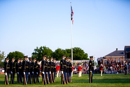 Soldiers in dark ceremonial uniforms are facing to the right while standing in rows with brown rifles at their sides. There is a white flagpole in the middle of the picture that has the US flag flying.