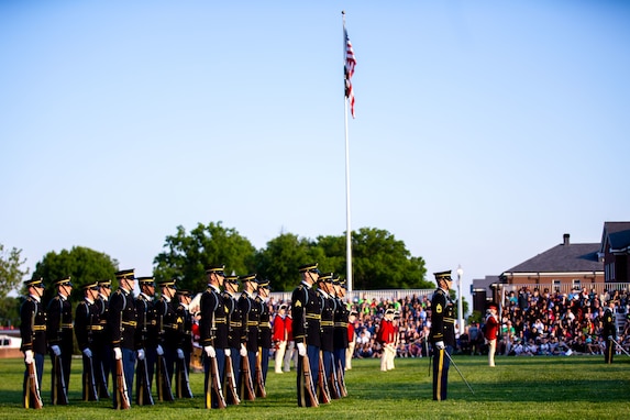 Soldiers in dark ceremonial uniforms are facing to the right while standing in rows with brown rifles at their sides. There is a white flagpole in the middle of the picture that has the US flag flying.