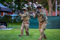 Army soldiers wearing green camouflage, helmets and dark goggles are walking forward with black rifles aimed in front of them. They are on a green lawn