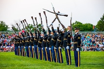 Soldiers dressed in dark Army ceremonial uniforms are standing in a straight row tossing brown rifles into the air one by one. They are on a green lawn and there is a crowd seated on bleachers in the background.